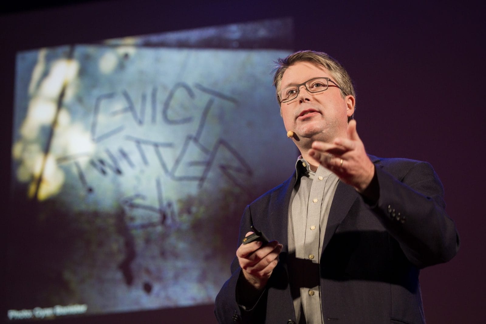 Dave Troy speaking on stage, gesturing, beside a projected slide showing graffiti that reads “EVICT TWITTER”